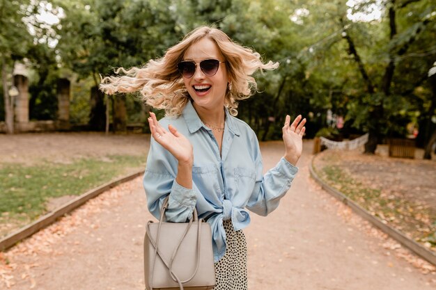 Atractiva rubia sonriente mujer sincera agitando el pelo largo divirtiéndose caminando en el parque en traje de verano