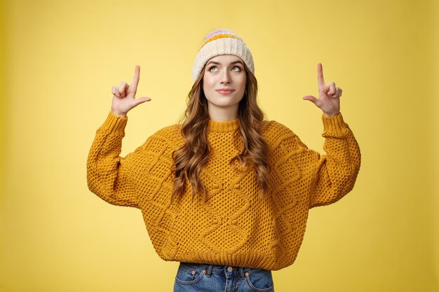 Atractiva soñadora elegante universitaria con sombrero suéter cálido mirando hacia arriba apuntando hacia arriba sonriendo deli ...