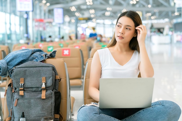 Atractiva tela casual femenina asiática con mochila viajero relajarse sentarse esperar en el asiento de distanciamiento social salida aeropuerto terminal mano usar computadora portátil seguridad concepto de viaje