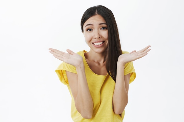 Atractiva tonta encantadora mujer de cabello oscuro con piel bronceada y sin maquillaje encogiéndose de hombros con las manos cerca de los hombros sonriendo ampliamente