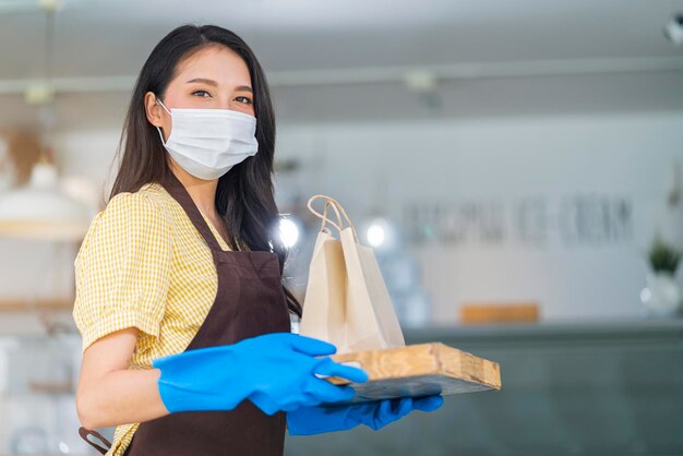 Atractiva trabajadora asiática de café usa mascarilla y guantes dando una bolsa de comida para llevar a la camarera con pedido para llevar de pie en la entrada de la puerta principal de la cafetería nuevo estilo de vida normal