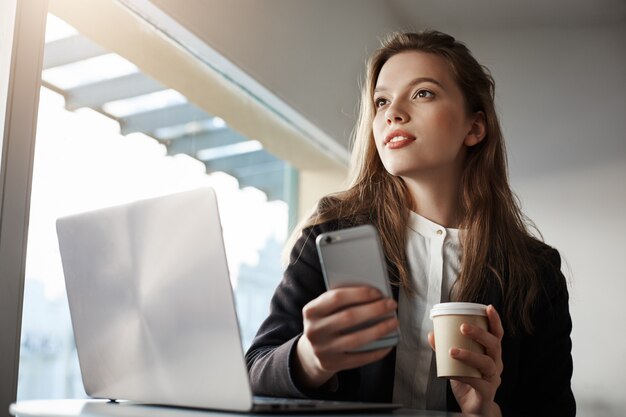 Atractiva trabajadora de oficina, sentada en la cafetería con una computadora portátil, tomando café y sosteniendo un teléfono inteligente, soñando mientras mira a un lado