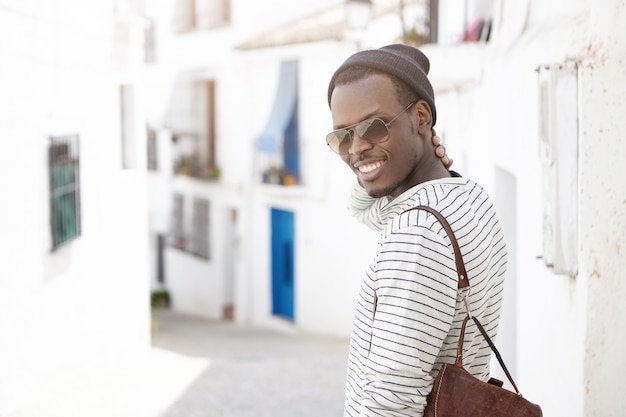 Atractivo y alegre turista europeo negro en tonos y sombrero caminando en las calles de una ciudad extranjera mientras pasa vacaciones en el extranjero. Concepto de personas, estilo de vida, viajes, aventura, turismo y vacaciones