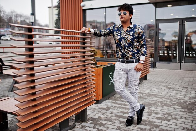 Atractivo y apuesto hombre indio elegante vestido con camisa y gafas de sol parado en la terraza del moderno café salón en su descanso de trabajo