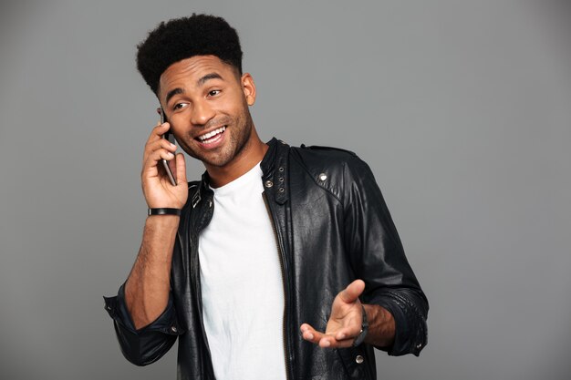 Atractivo chico africano feliz con elegante corte de pelo hablando por teléfono inteligente, mirando a un lado