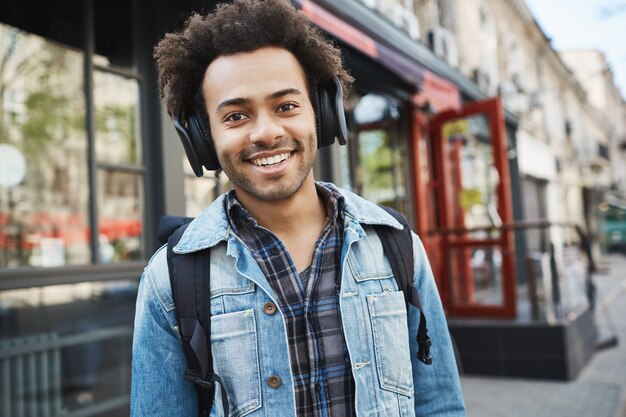 Atractivo chico sonriente de piel oscura con cerdas, escuchando música mientras camina por la calle, de buen humor