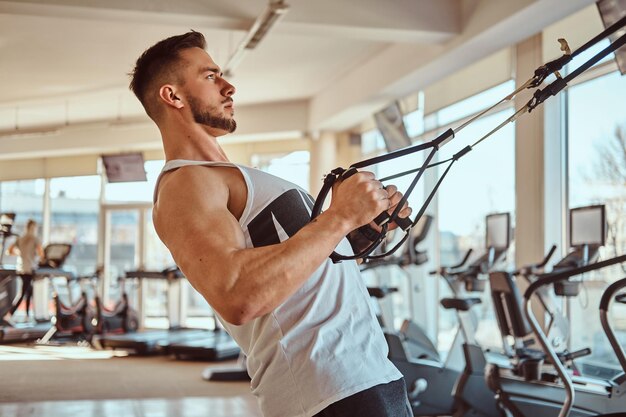 Atractivo culturista poderoso está haciendo ejercicios en aparatos de entrenamiento en el gimnasio soleado.