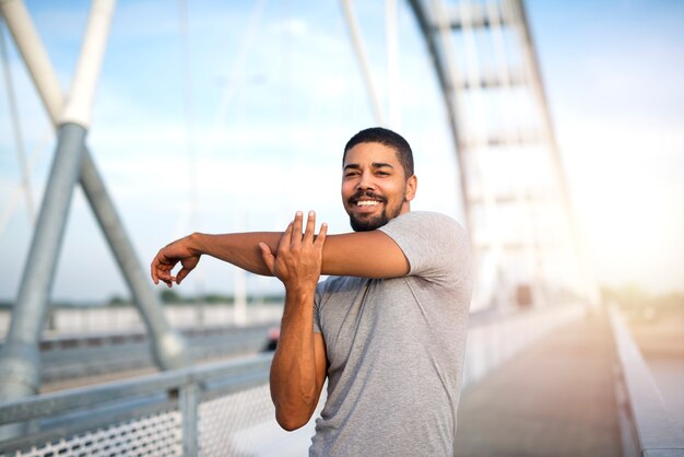 Atractivo deportista sonriente calentando su cuerpo para un entrenamiento al aire libre