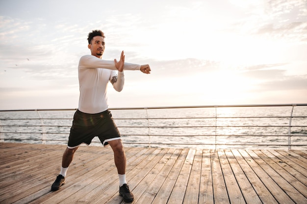 Atractivo y encantador deportista de piel oscura con pantalones cortos negros y camiseta blanca de manga larga hace ejercicios de arca, se estira y trabaja cerca del mar