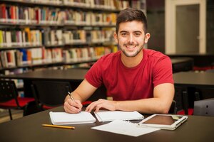 Foto gratuita atractivo estudiante universitario masculino haciendo algunos deberes en la biblioteca de la escuela y sonriendo
