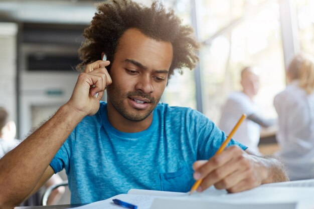 Atractivo estudiante universitario de piel oscura y barbudo que trabaja en el papel del curso en el espacio de coworking, tomando notas en un libro de texto con lápiz mientras habla con su supervisor de investigación por teléfono móvil. Efecto de película