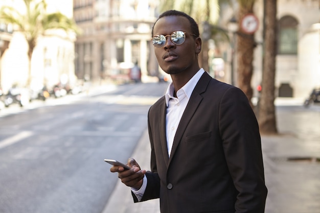 Atractivo y exitoso empresario afroamericano con traje formal negro y gafas de sol con lentes de espejo de pie en la calle con teléfono inteligente, llamando a un taxi, mirando hacia adelante con impaciencia