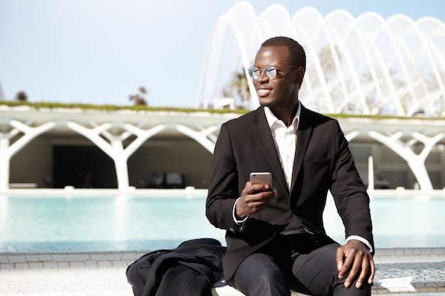 Atractivo gerente afroamericano con elegantes vestimentas formales y tonos usando un teléfono móvil, sentado en un banco en un entorno urbano mientras espera a almorzar a sus colegas, sonriendo alegremente cuando los nota