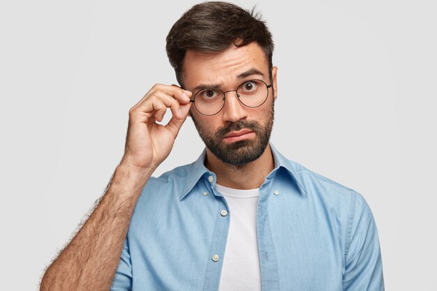 Atractivo hombre sin afeitar mira con curiosidad a través de gafas, mantiene la mano en el borde, vestido con una camisa de moda, posa contra la pared blanca. Joven escucha algo interesante con asombro