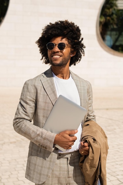 Atractivo hombre afroamericano al aire libre. Hombre de traje con barba sosteniendo portátil, caminando. Mirando a la cámara. Retrato, concepto de vida de la ciudad