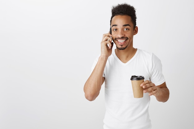Atractivo hombre afroamericano sonriente hablando por teléfono y bebiendo café para llevar