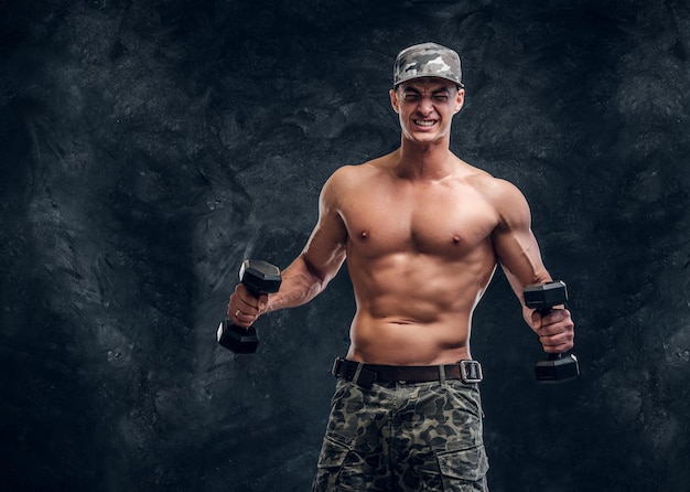 Atractivo hombre sin camisa con gorra está haciendo ejercicios con pesas sobre fondo oscuro.