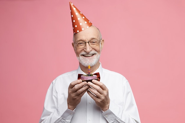 Atractivo hombre caucásico jubilado feliz con pajarita, gafas y sombrero de cono celebrando su 80 aniversario, posando aislado con pastel de cumpleaños en sus manos, yendo a soplar velas y pedir un deseo