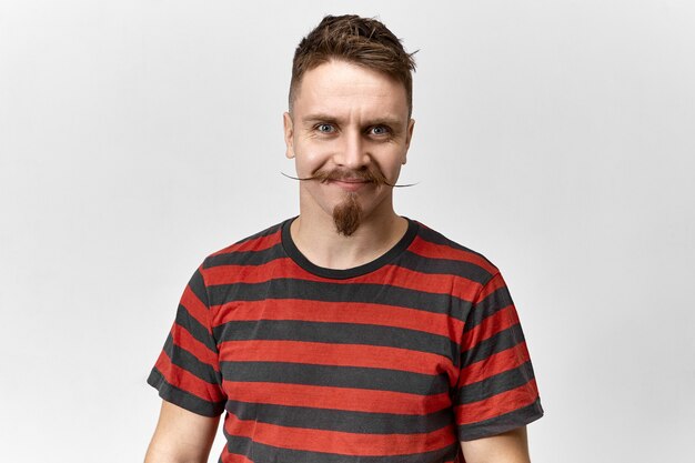 Atractivo hombre caucásico morena de ojos azules vistiendo elegante camiseta con rayas rojas y negras de buen humor. Chico hipster guapo positivo con bigote de manillar y perilla posando en el estudio