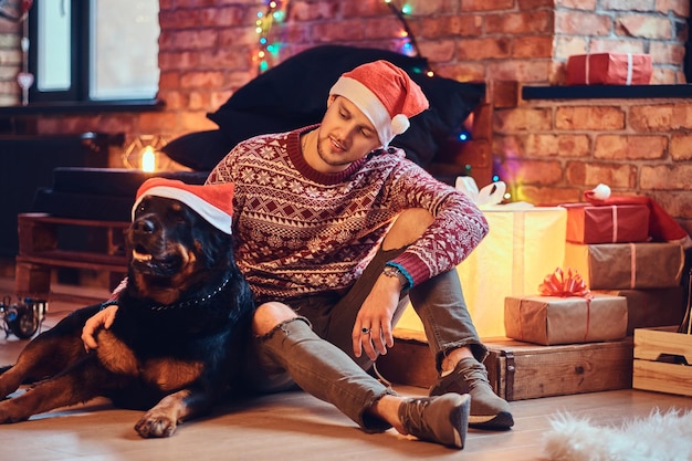 Atractivo hombre hipster barbudo se sienta en el suelo con su perro Rottweiler en una habitación con decoración navideña.