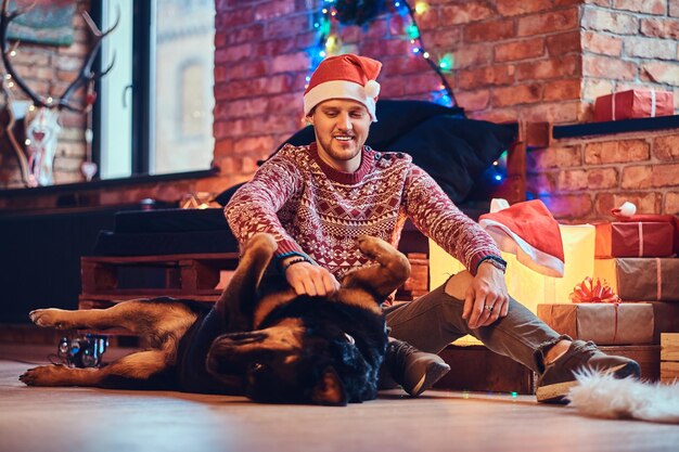 Atractivo hombre hipster barbudo se sienta en el suelo con su perro Rottweiler en una habitación con decoración navideña.