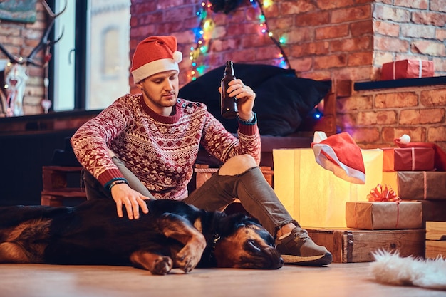 Atractivo hombre hipster barbudo se sienta en el suelo con su perro Rottweiler en una habitación con decoración navideña.