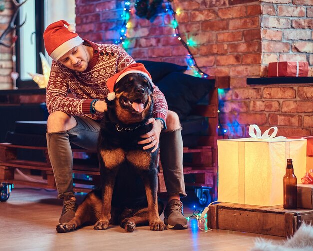 Atractivo hombre hipster barbudo con su perro Rottweiler en una habitación con decoración navideña.