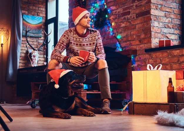 Atractivo hombre hipster barbudo con su perro Rottweiler en una habitación con decoración navideña.