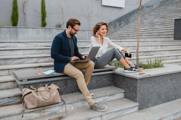 Atractivo hombre y mujer sentados en las escaleras en el centro urbano de la ciudad, hablando por auriculares inalámbricos manos libres
