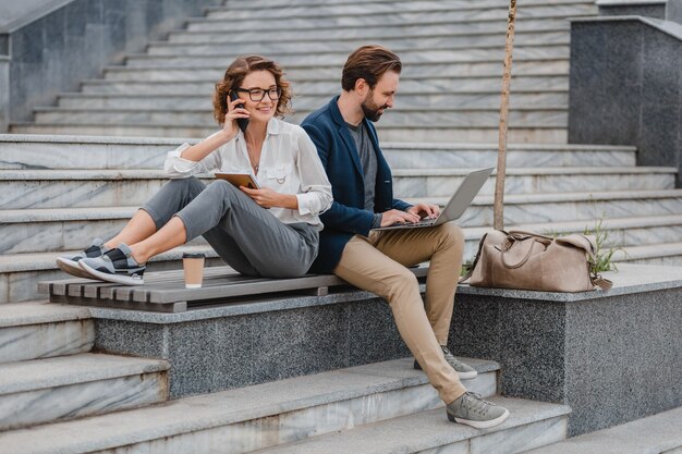 Atractivo hombre y mujer sentados en las escaleras en el centro urbano de la ciudad, trabajando juntos en la computadora portátil