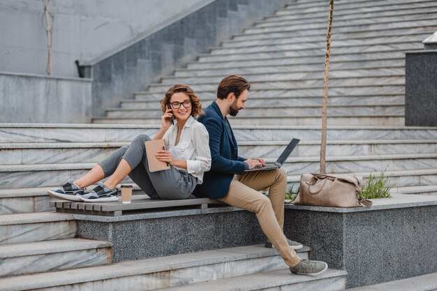 Atractivo hombre y mujer sentados en las escaleras en el centro urbano de la ciudad, trabajando juntos en la computadora portátil