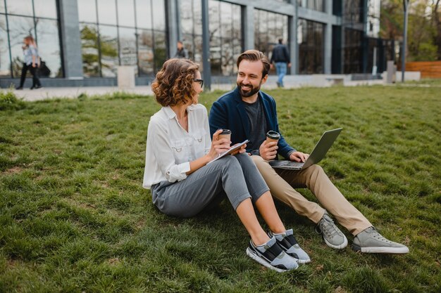 Atractivo hombre y mujer sonriente hablando sentados en el césped en el parque urbano, tomando notas