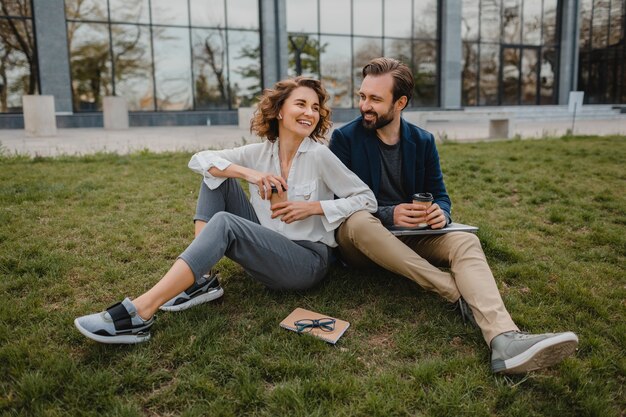 Atractivo hombre y mujer sonriente hablando sentados en el césped en el parque urbano, tomando notas