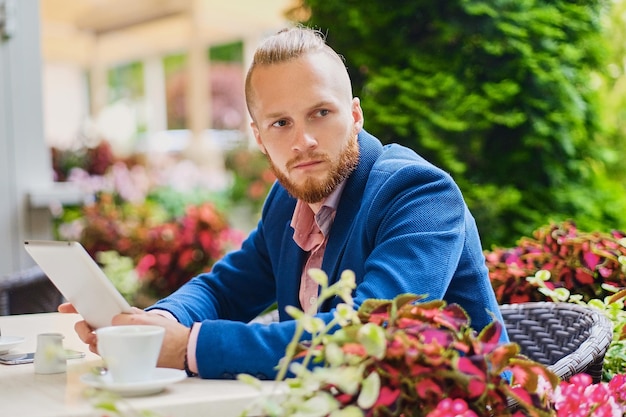 Atractivo hombre pelirrojo barbudo con una camisa rosa y una chaqueta azul se sienta en la mesa de un café y usa una tableta.