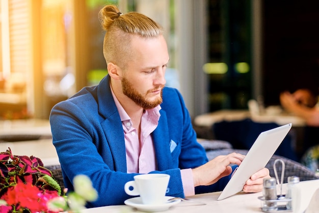 Atractivo hombre pelirrojo barbudo con una camisa rosa y una chaqueta azul se sienta en la mesa de un café y usa una tableta.