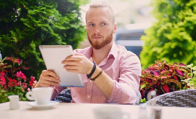 Atractivo hombre pelirrojo barbudo con una camisa rosa se sienta en la mesa de un café y usa una tableta.