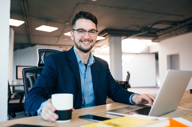 Atractivo hombre de pelo oscuro está trabajando en la mesa en la oficina. Viste camisa azul con chaqueta negra. Está tomando una taza de café y sonriendo a la cámara.