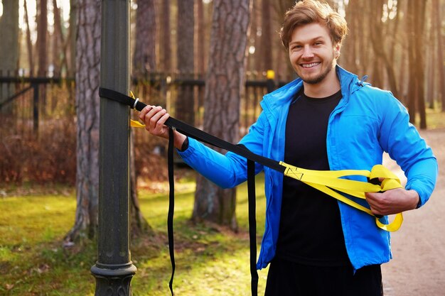 Atractivo hombre positivo con un impermeable azul haciendo ejercicio en un parque con tiras de fitness trx.