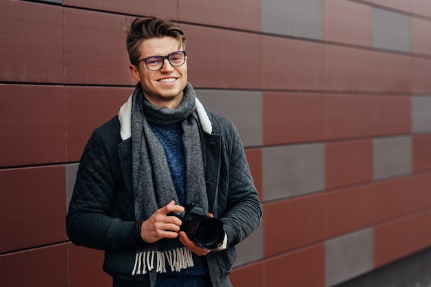 Atractivo hombre sonriente en anteojos tiene cámara profesional al aire libre contra la pared.