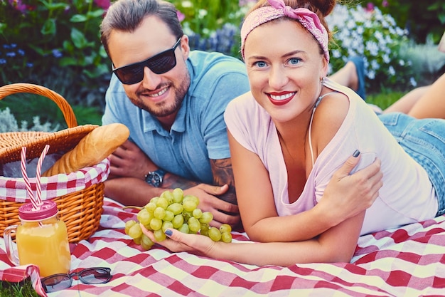 Atractivo hombre tatuado, barbudo y una mujer pelirroja acostada sobre una manta en un césped en un parque. tiempo de pícnic.