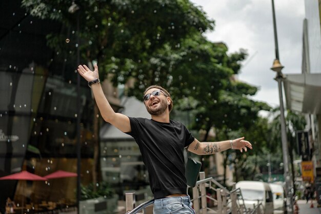 Atractivo hombre tatuado feliz sonriendo en la calle en Kuala Lumpur. Hombres urbanos, estilo de ropa. Las burbujas de jabón vuelan en el aire.