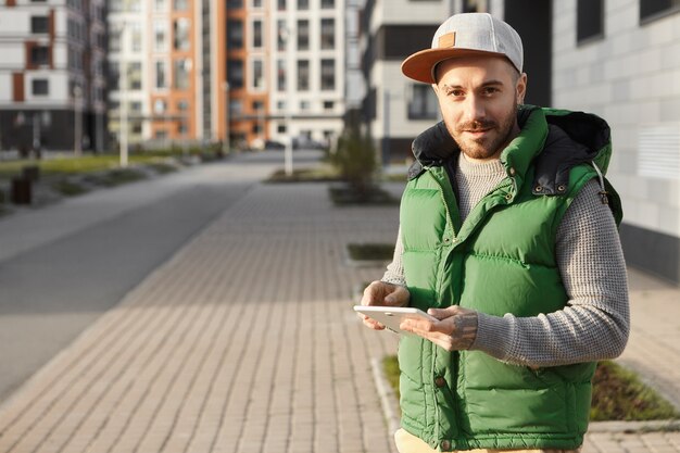 Atractivo joven sin afeitar en snapback permaneciendo siempre conectado, enviando mensajes a amigos en línea usando el teléfono afuera en la calle de la ciudad. Chico lindo hipster escribiendo mensaje en el panel táctil