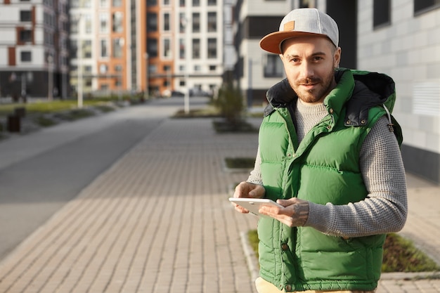 Atractivo joven sin afeitar en snapback permaneciendo siempre conectado, enviando mensajes a amigos en línea usando el teléfono afuera en la calle de la ciudad. Chico lindo hipster escribiendo mensaje en el panel táctil