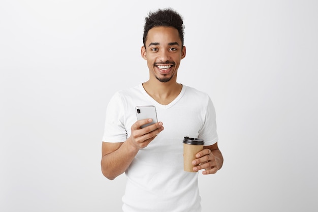 Atractivo joven afroamericano mirando complacido, sosteniendo el teléfono móvil y bebiendo café para llevar