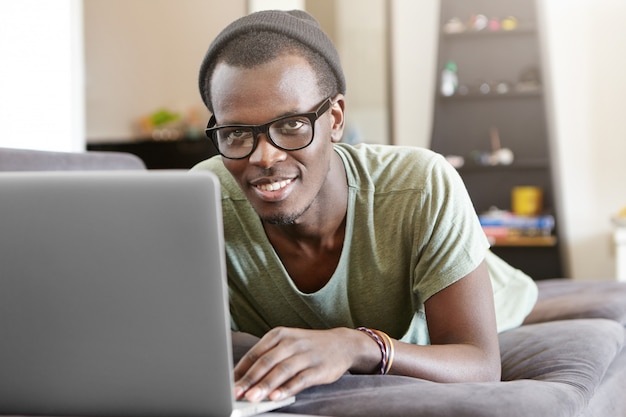 Atractivo joven afroamericano relajante en casa con pc portátil, acostado en el sofá gris solo después del trabajo. Hipster negro con videollamada