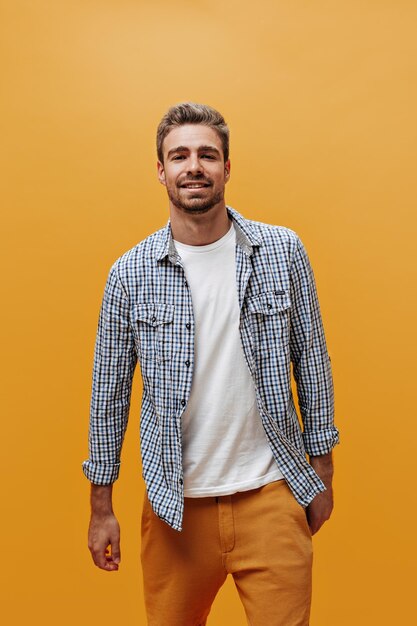 Atractivo joven barbudo con camisa a cuadros azul y camiseta blanca se ve seguro de sí mismo y posa sobre fondo naranja