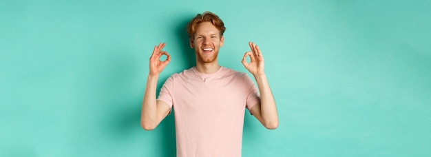 Foto gratuita atractivo joven en camiseta sonriendo satisfecho asintiendo con aprobación y mostrando el signo ok aprobar y agr