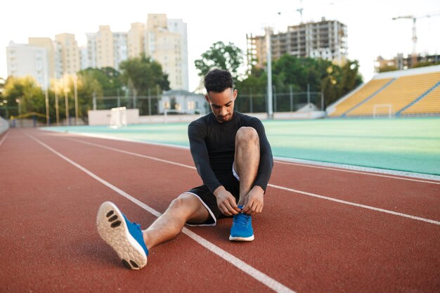 Atractivo joven deportista afroamericano atando cordones de zapatos en zapatillas de deporte en la pista de carreras en el estadio de la ciudad