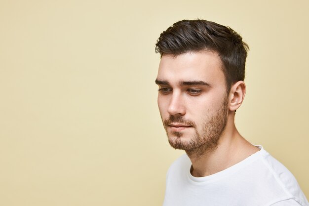 Atractivo joven europeo con cerdas mirando hacia abajo con sonrisa tímida posando en camiseta blanca contra la pared en blanco con espacio de copia para su información publicitaria