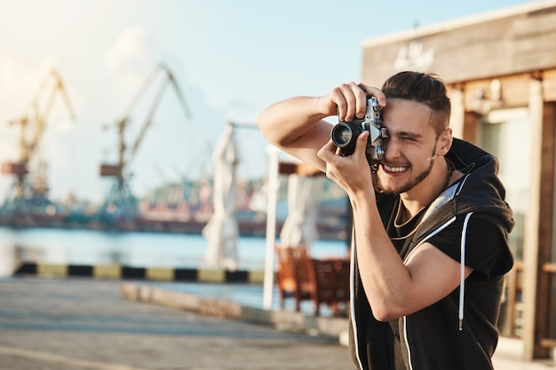 Atractivo joven fotógrafo masculino caminando por el puerto, haciendo fotos de yates y personas geniales, mirando a través de la cámara enfocada en una gran toma, con talento para el fotoperiodismo